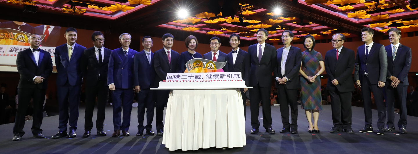 Thirteen people in suits pose behind a BreadTalk cake on stage.